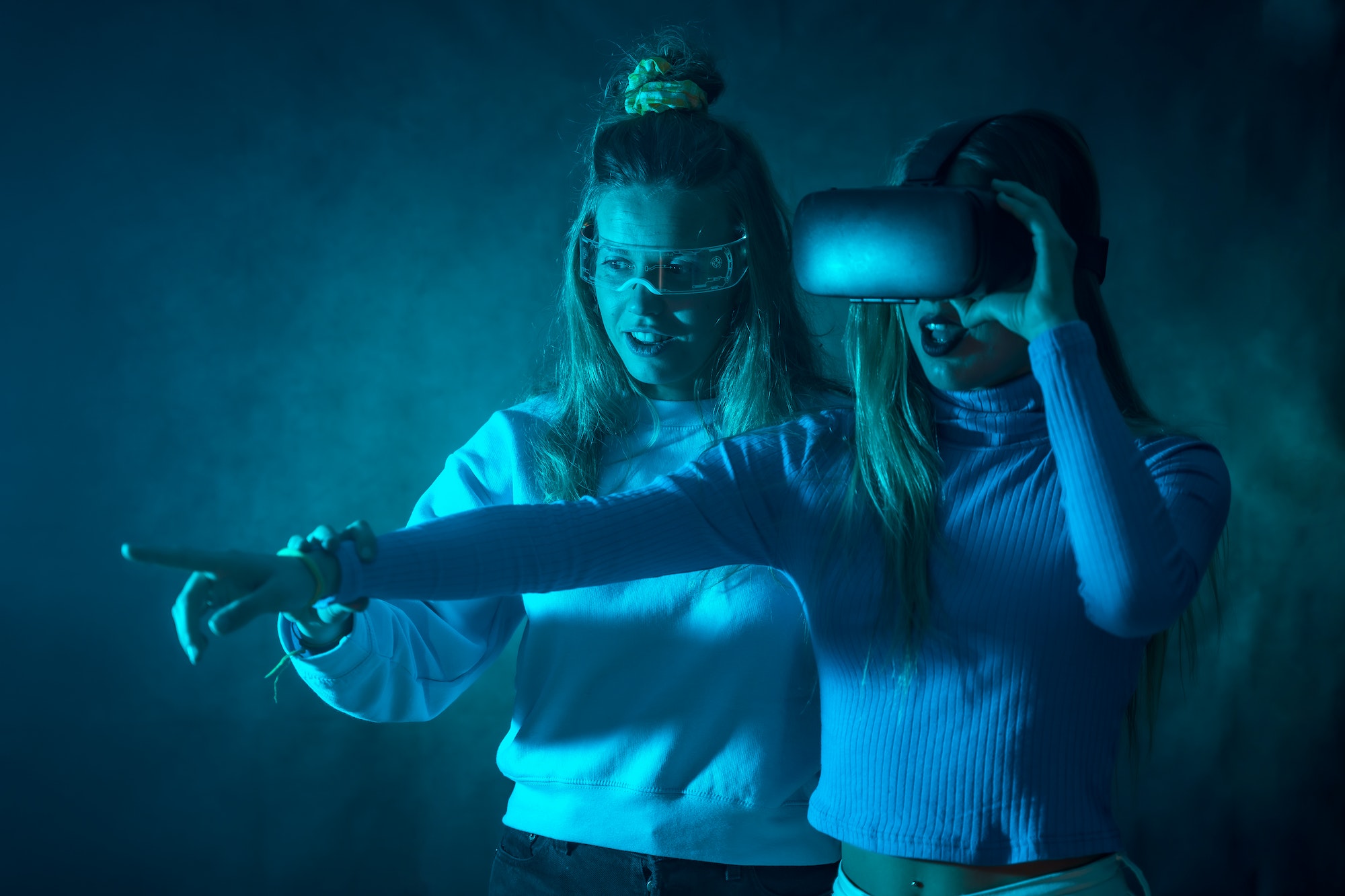 Two futuristic women in vr glasses, pointing virtual reality things, blue background